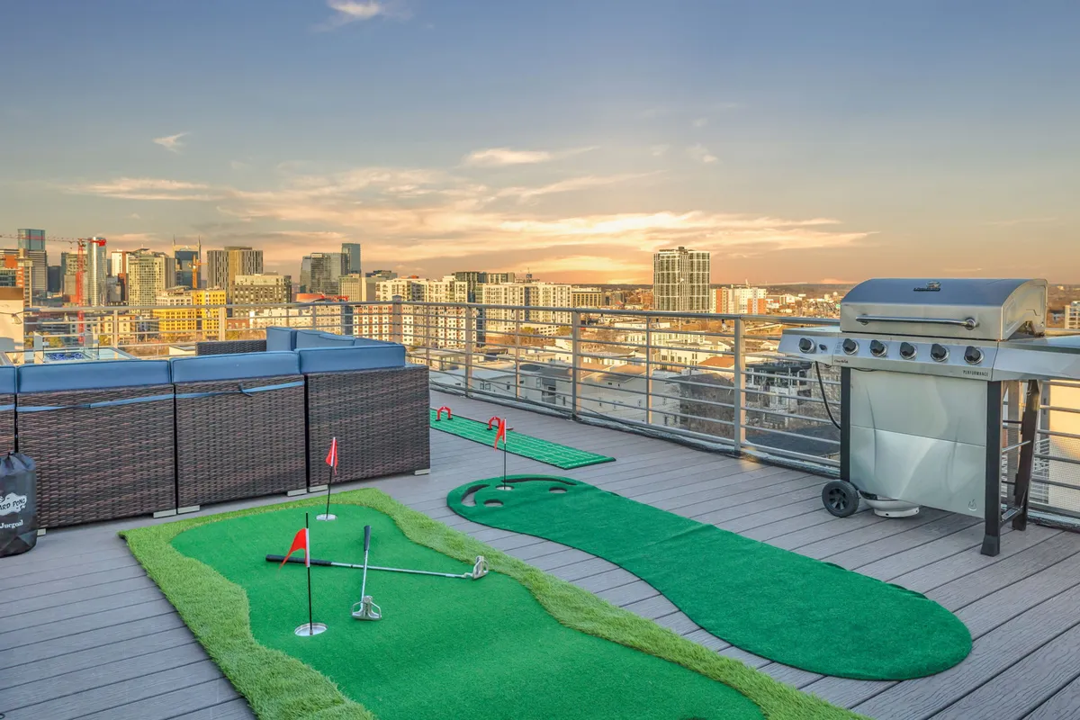 A well-appointed rooftop deck featuring a putting green for mini-golf, a grill for outdoor feasts, modern seating, and the city skyline glowing in the background. The space is clearly designed for both play and leisure, with sunset hues enhancing the ambiance.