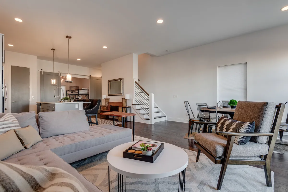 A warm, inviting living and dining area featuring a plush sofa, coffee table, formal dining table, and a staircase leading to other areas of the home. The layout emphasizes social interaction and comfortable group gatherings.