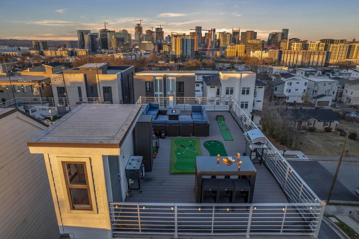 A spacious rooftop patio outfitted with modern outdoor furniture, a putting green, and an al fresco dining area. The Nashville skyline creates a dramatic backdrop while the rooftop is set up for both relaxation and playful activities, with string lights adding a festive touch.