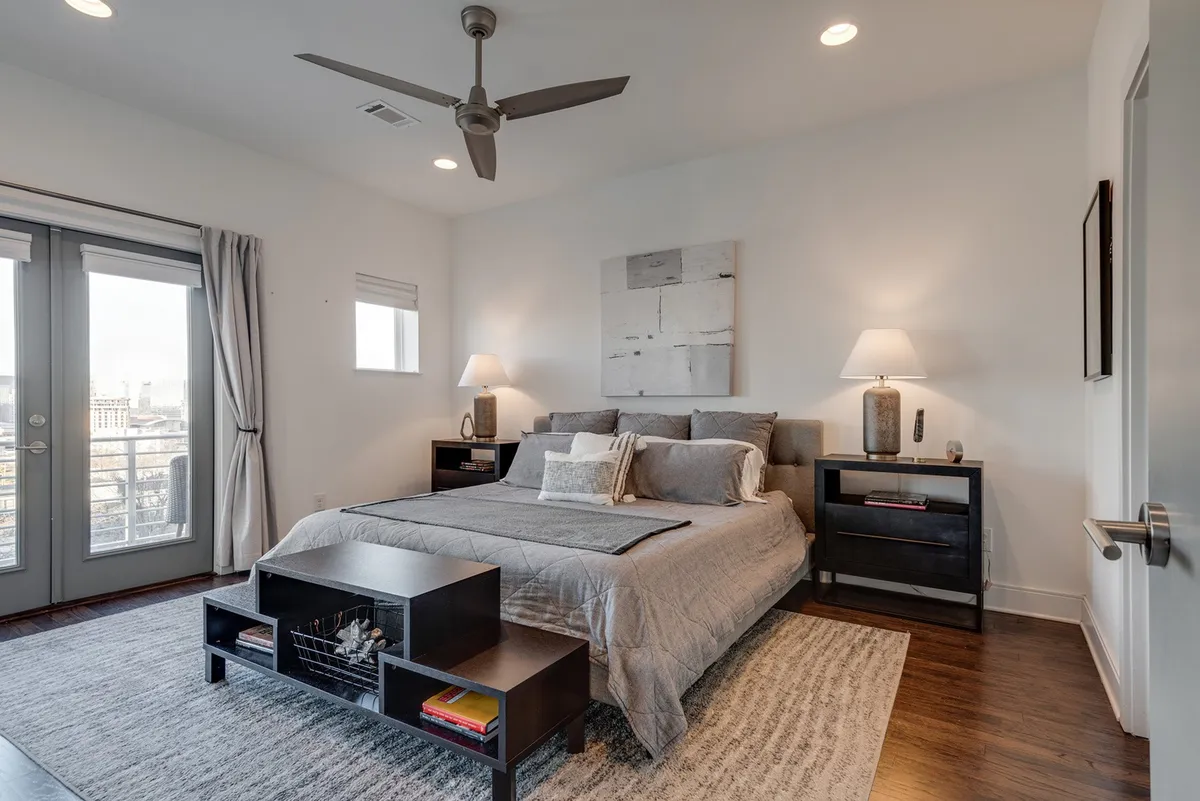 This image displays a tranquil bedroom with a gray bed, matching nightstands with lamps, wall art, and sliding doors that open to an outdoor space. The abundance of natural light and serene decor create a sense of restful privacy and comfort.