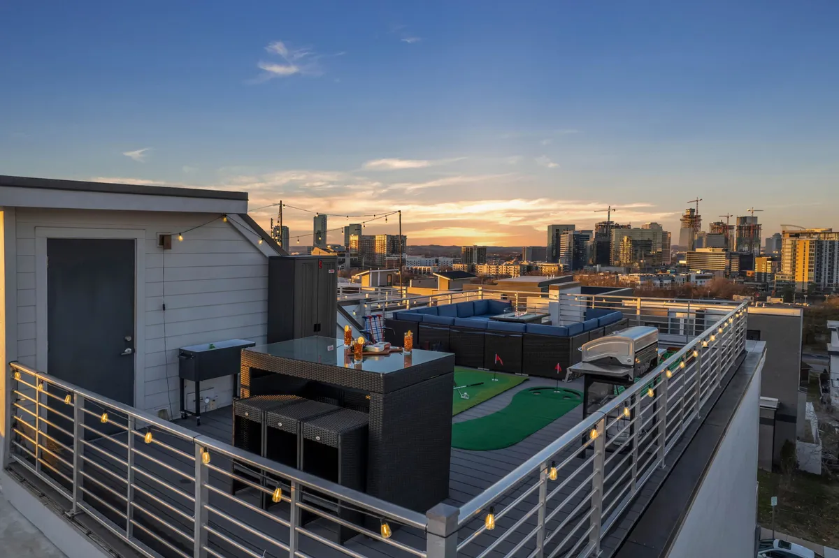 A vibrant rooftop scene featuring an inviting seating area, a fire pit, a mini-golf putting green, and sweeping views of the Nashville city skyline at sunset. String lights add a festive atmosphere, and the arrangement is clearly set for socializing, with spaces for both relaxation and playful competition.