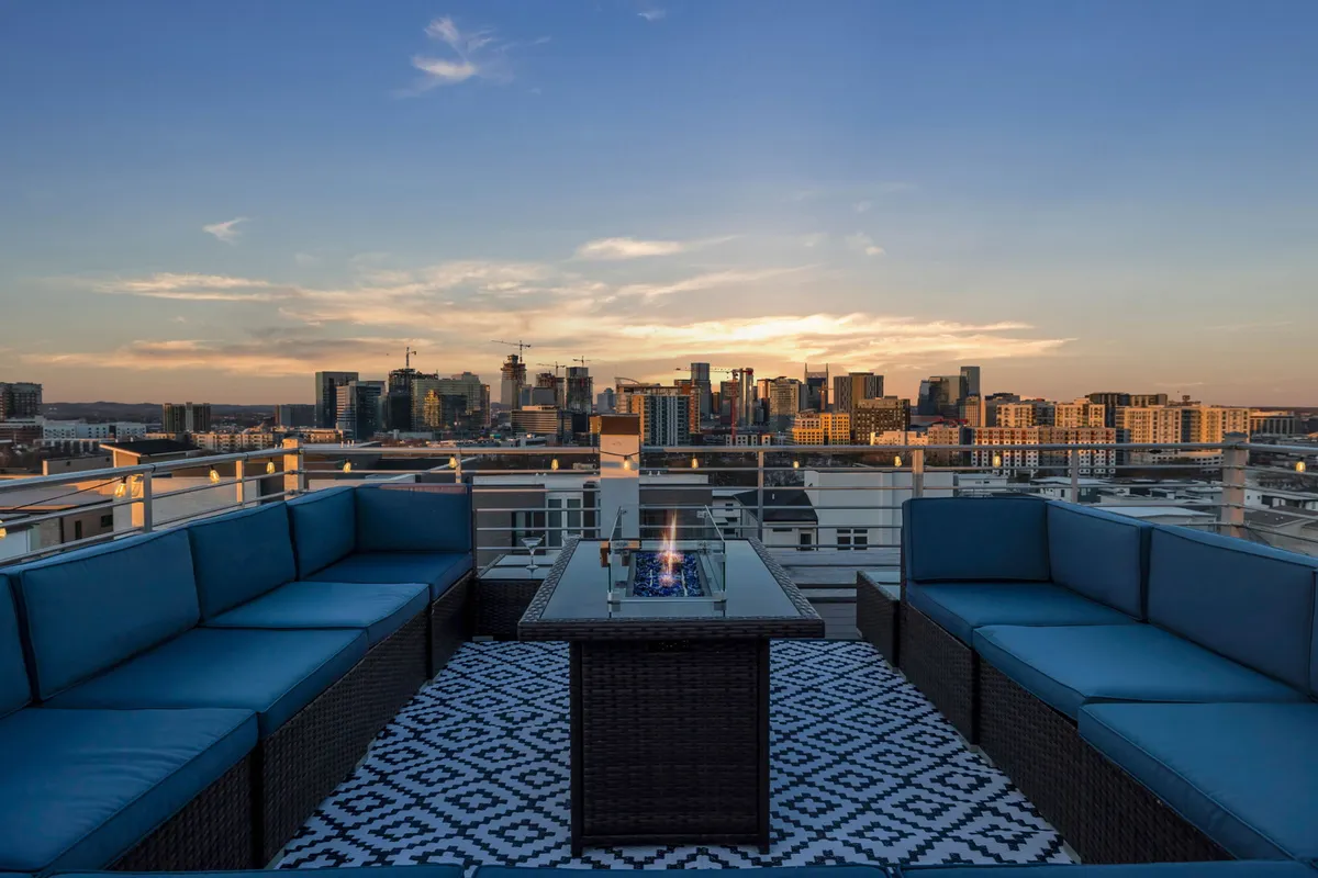 A rooftop seating area featuring a fire pit, comfortable outdoor furniture, and panoramic views of the city skyline at sunset. Groups are pictured socializing and relaxing as city lights begin to twinkle in the background, capturing a cozy yet lively atmosphere.