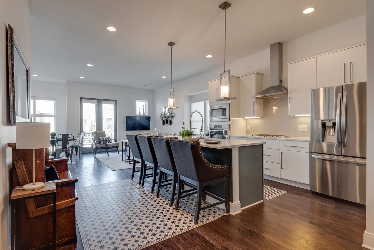 A bright, modern kitchen with an island countertop, stainless steel appliances, dining chairs, a decorative rug, and large windows that let in plenty of natural light. The space suggests a welcoming environment ideal for both meal preparation and casual gatherings.