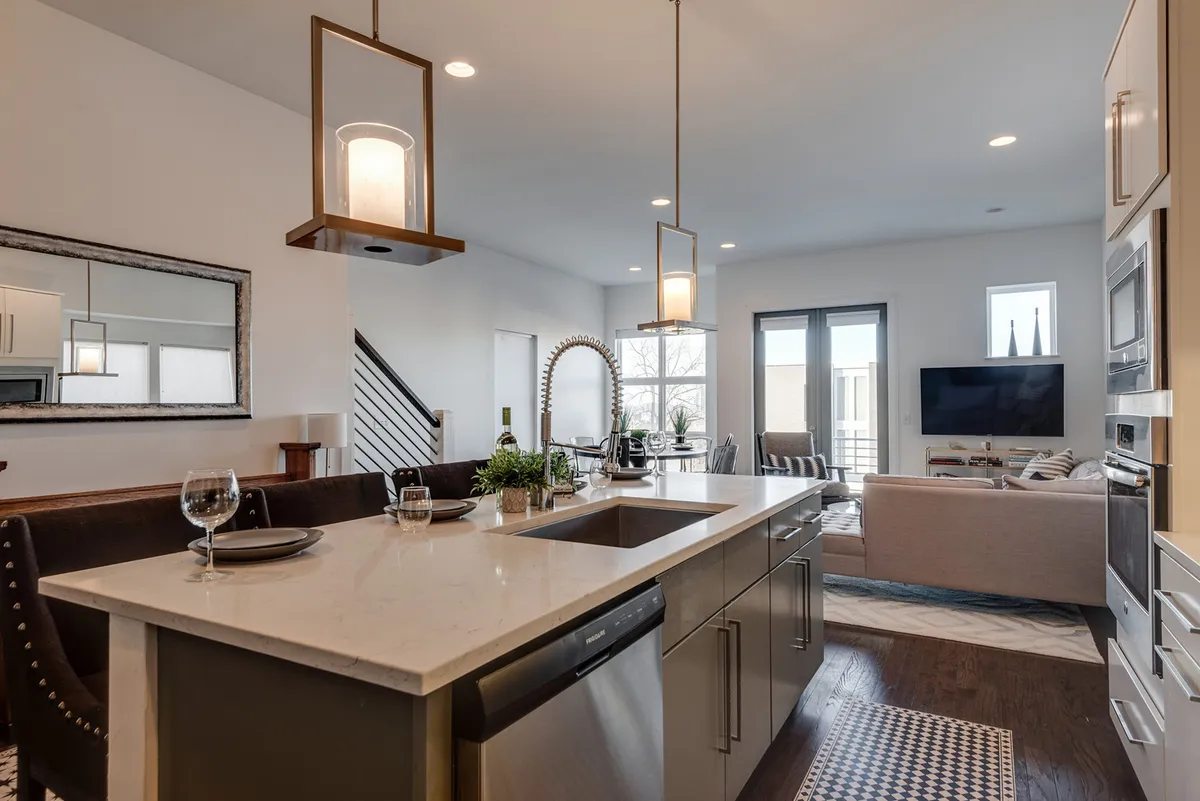 An open-plan interior with a kitchen island illuminated by pendant lights, a TV area with comfortable seating, and an adjacent dining space. The layout is designed for socializing, cooking, and relaxing, with stylish decor and abundant light.