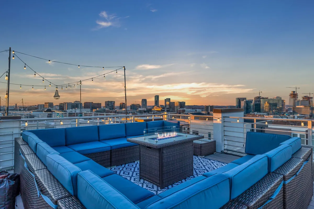 A cozy, stylish rooftop lounge area where a blue sectional sofa surrounds a central fire pit. Hanging string lights and the illuminated city skyline create an intimate, vibrant setting perfect for sunset toasts, stargazing, or group celebrations.